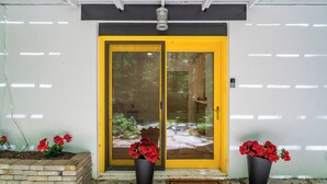 A yellow door with a glass panel, two potted red flowers, and a welcome mat.