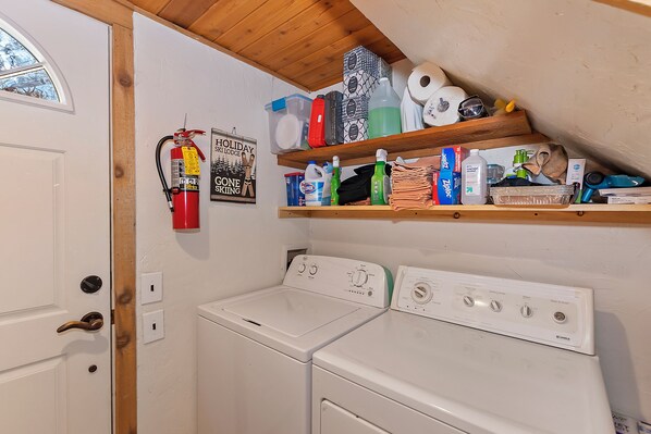 A laundry area with two white washers and dryers, a fire extinguisher, and shelves above holding various cleaning supplies and toiletries.