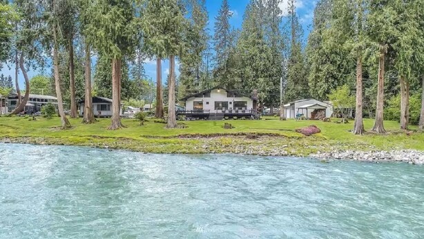 Una propiedad a orillas del río con una casa rodeada de altos árboles, una zona cubierta de césped y vistas despejadas al río.