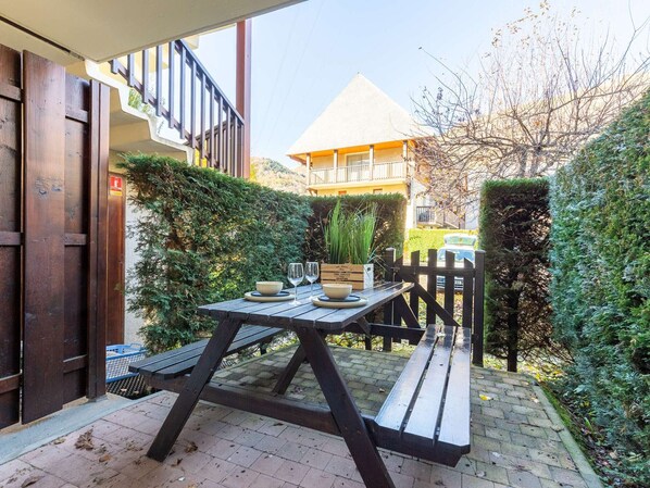 An outdoor patio with a wooden table and benches, surrounded by greenery and a fence.