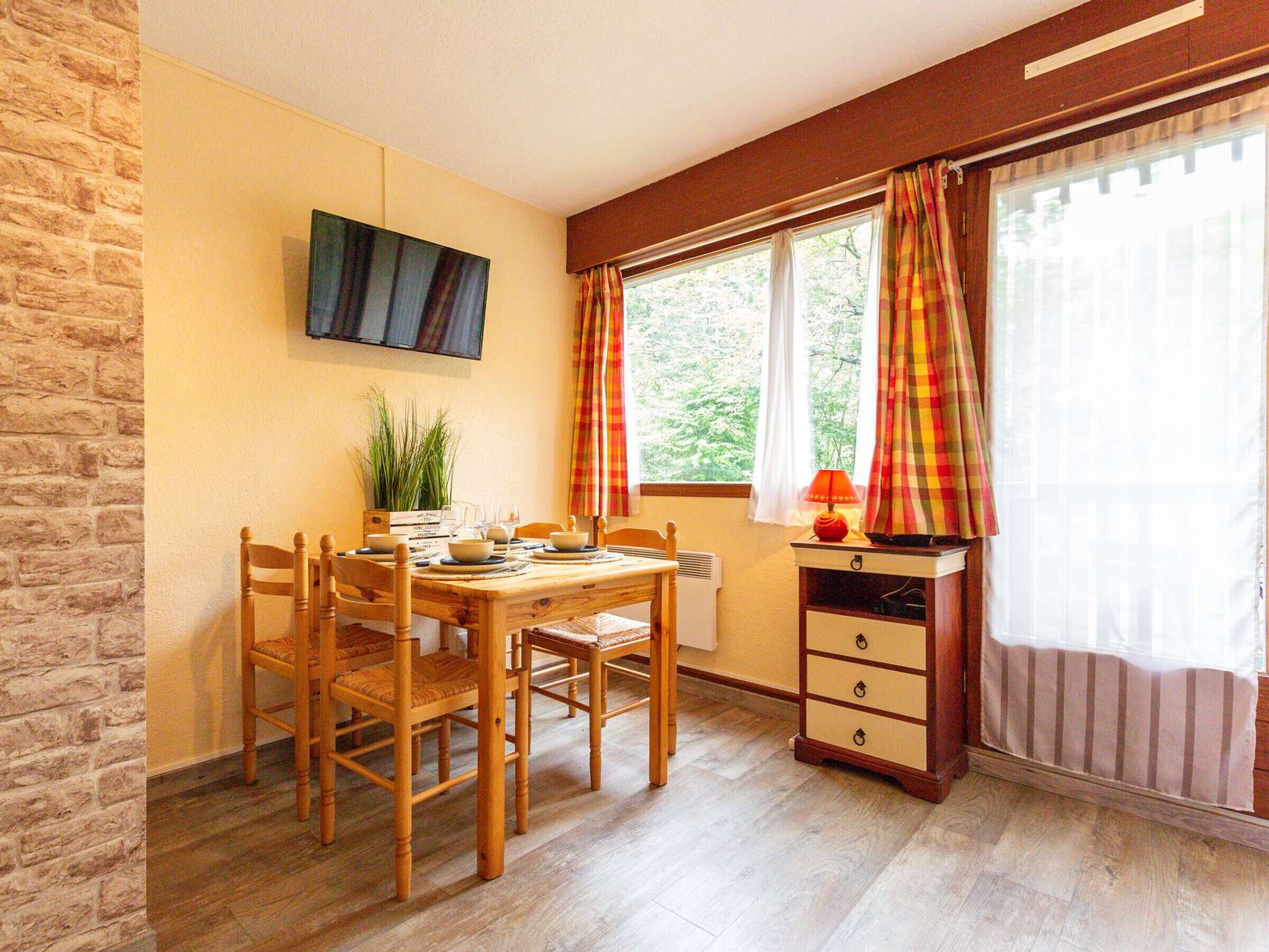 Un comedor con una mesa y sillas de madera, un televisor montado en la pared y una ventana con cortinas.