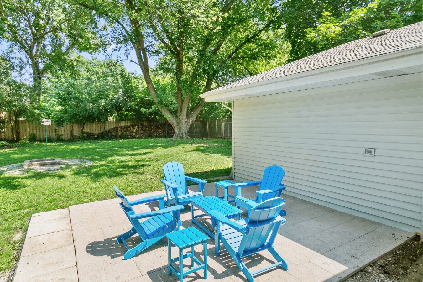 Un patio con sillas Adirondack azules y una pequeña mesa, un edificio blanco y un césped con árboles.