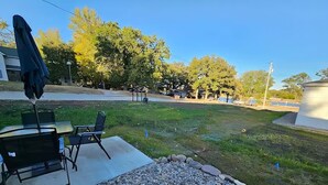 Uma área de pátio com mesa e cadeiras, um pequeno jardim com pedras e vista para árvores e um edifício ao fundo.