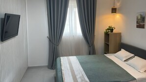 A modern hotel room with a bed, curtains, a TV mounted on the wall, a bedside table with a plant, and a wall-mounted light fixture. - Charming 3-bedroom house in Figueira da Foz, Serra da Boa Viagem (Quiaios)