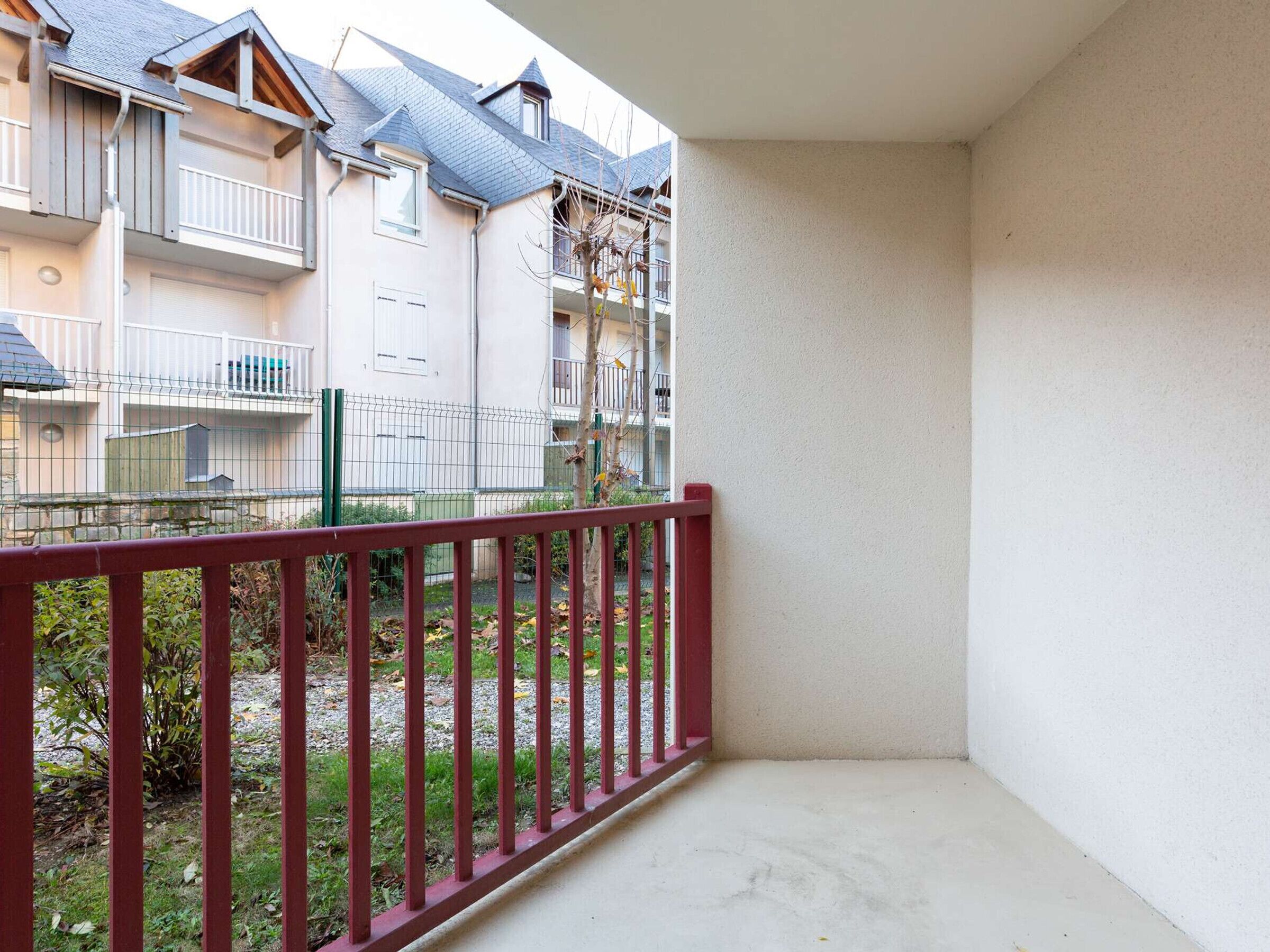Un balcón con barandilla roja, vista de un edificio residencial con varios balcones y un área verde con árboles.