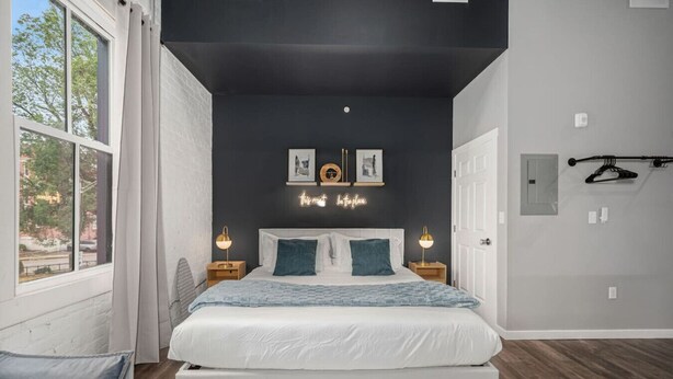 Ein modernes Schlafzimmer mit einem großen Bett, zwei Nachttischen mit Lampen, einem Neon-Schild an der Wand und Blick ins Freie durch ein Fenster.