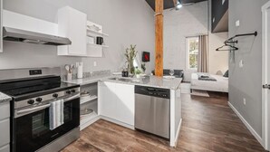Eine moderne Küche mit Einbauherd, Backofen und Geschirrspüler, einer Holzbalkendecke und Blick in einen Wohnbereich mit Sofa und Fenster.