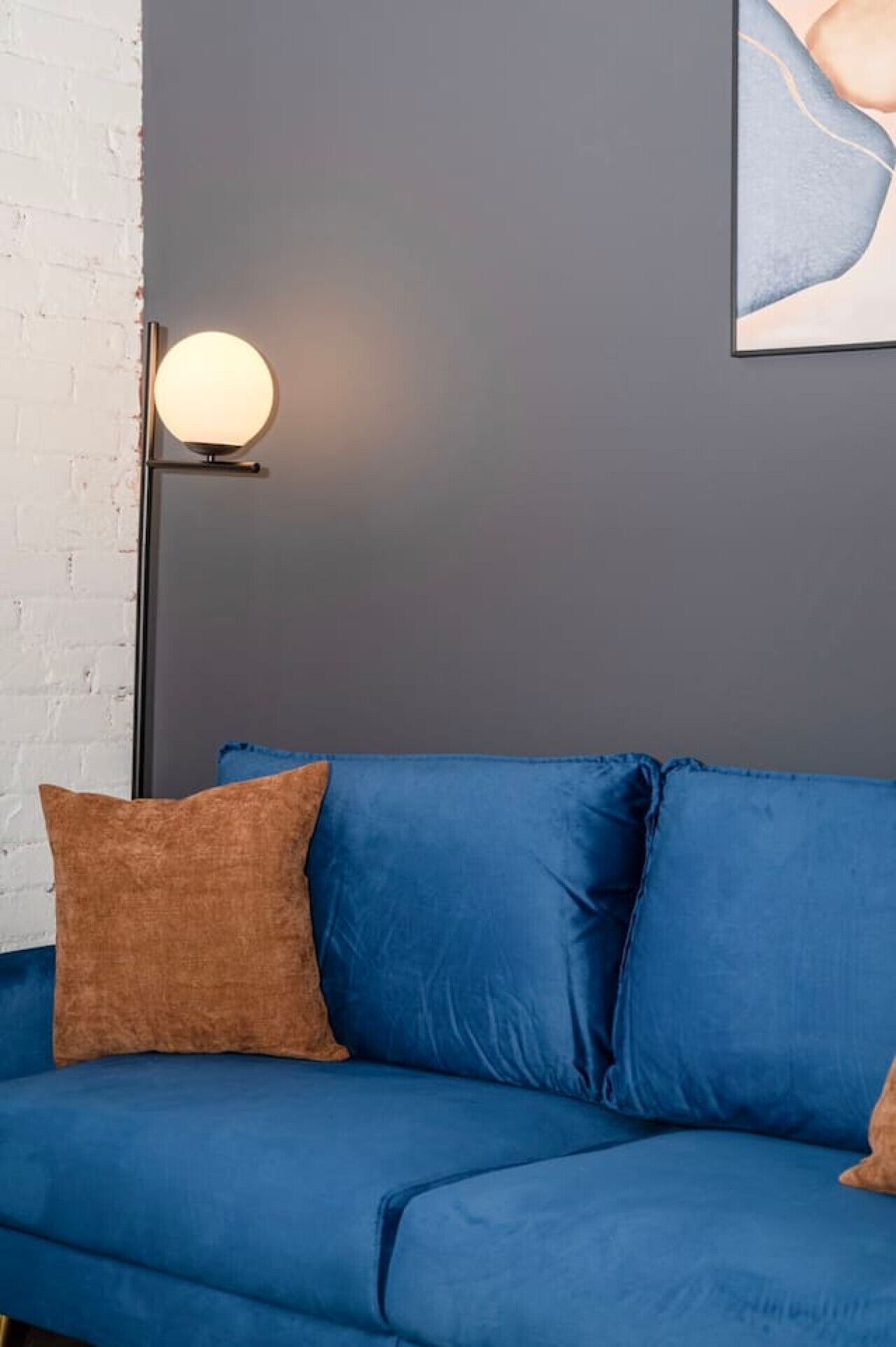 A blue sofa with two cushions, a floor lamp, and a white brick wall.