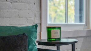 A room with a white brick wall, a window with a view of trees, a green cushion on a dark sofa, and a small table with a green candle.