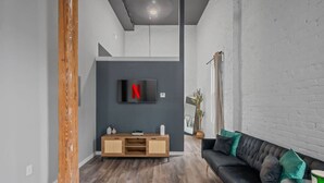 A modern living room with a dark grey accent wall, a wooden cabinet, a black leather sofa with green cushions, and a mounted TV displaying the Netflix logo.