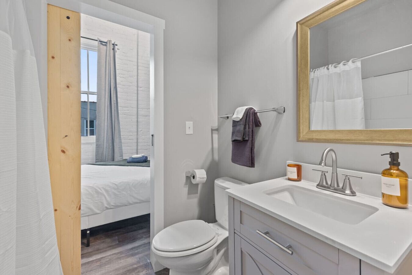 Ein modernes Badezimmer mit einem weißen Waschbecken, einem Spiegel und einer Toilette. Ein Fenster mit Vorhang ist vorhanden, und im angrenzenden Raum ist ein Bett zu sehen.