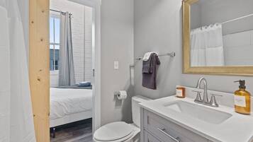 Ein modernes Badezimmer mit einem weißen Waschbecken, einem Spiegel und einer Toilette. Ein Fenster mit Vorhang ist vorhanden, und im angrenzenden Raum ist ein Bett zu sehen.