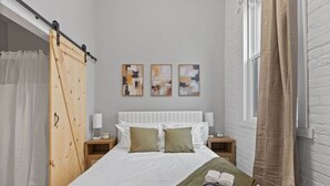 Una cama bien hecha con una manta verde y toallas enrolladas, un cabecero blanco y tres cuadros abstractos en la pared.