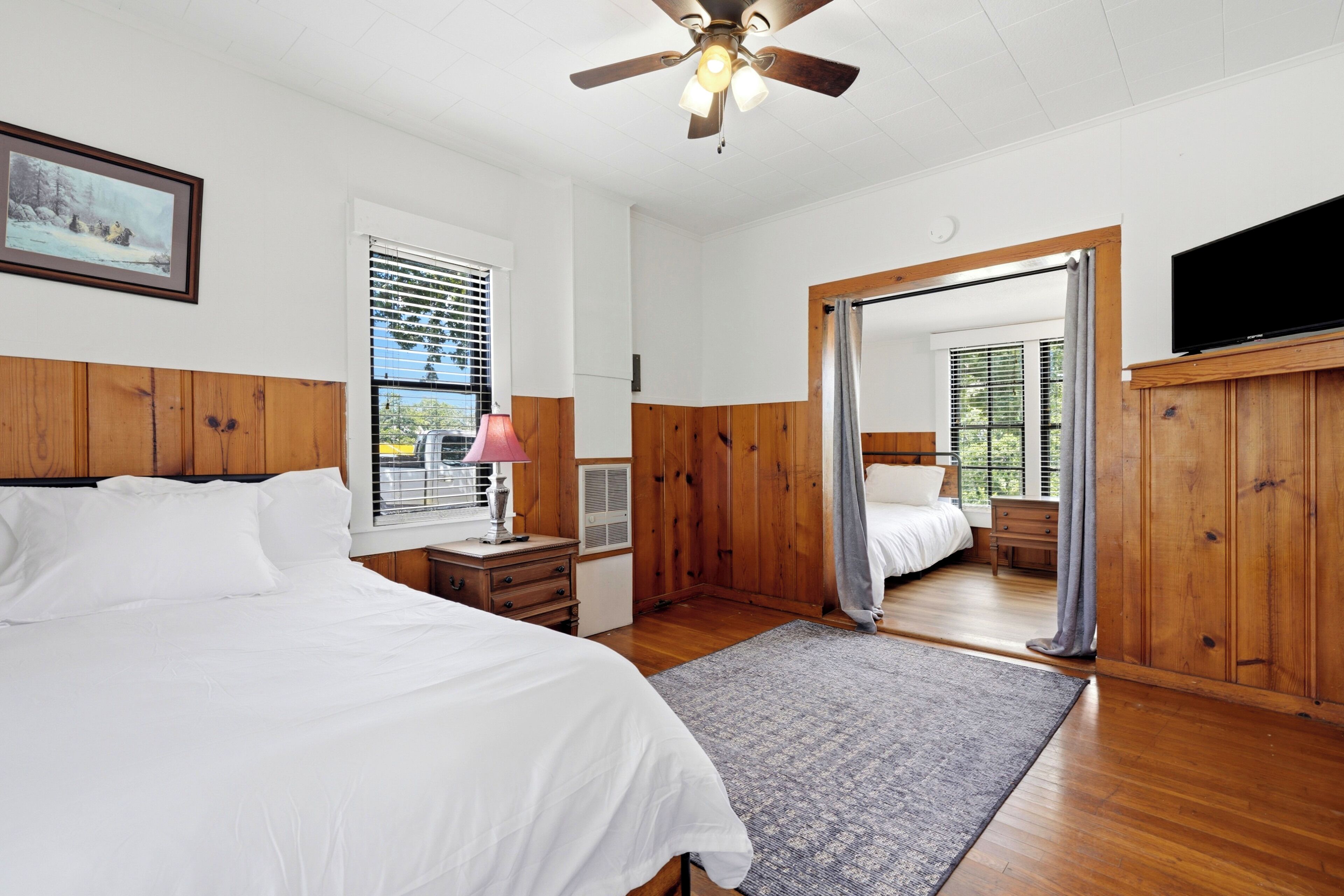 A bedroom with a bed, a desk, a lamp, a ceiling fan, and a television.