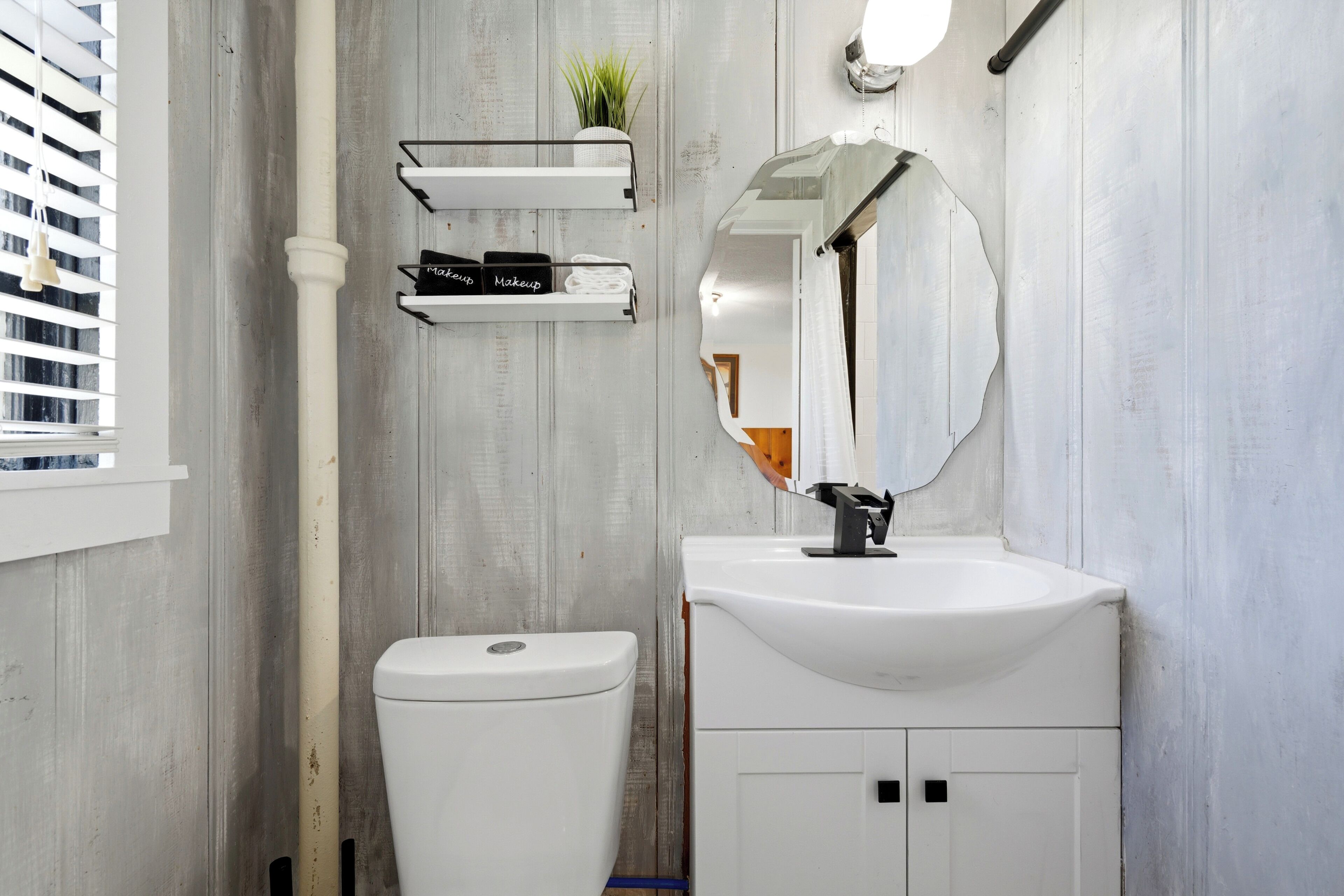 A bathroom with a white toilet, a sink with a mirror, and a shelf with makeup items.
