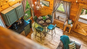 A cozy cabin interior with a dining table set for four, surrounded by green chairs. A chandelier hangs above the table, and a kitchen area is visible to the left.