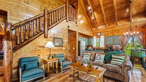 A wooden cabin interior with a staircase, a living room with a sofa and chairs, a kitchen area, and a dining table.