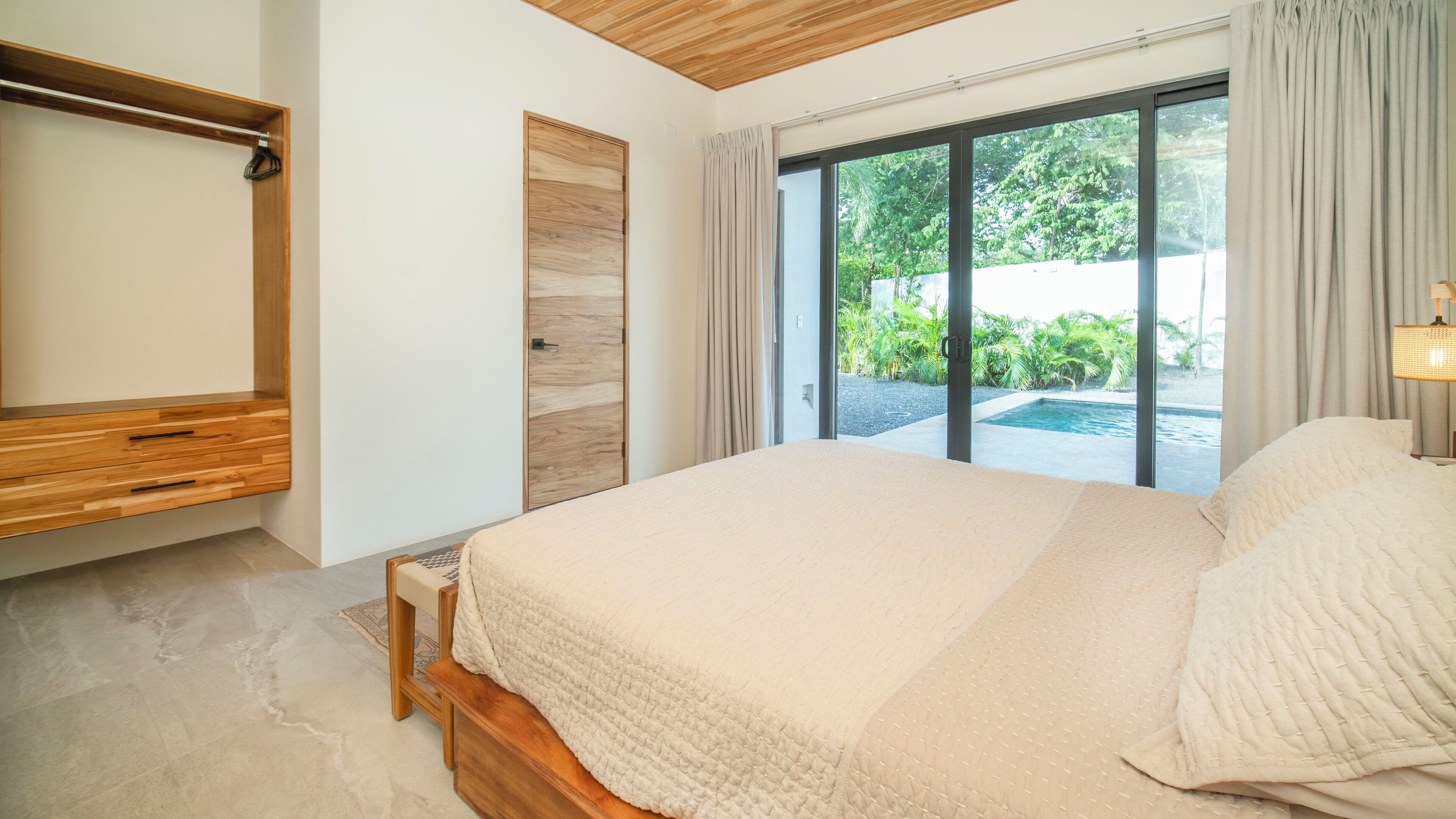 Un dormitorio con una cama grande, un armario de madera, un pequeño banco y vistas a una piscina y zonas verdes.