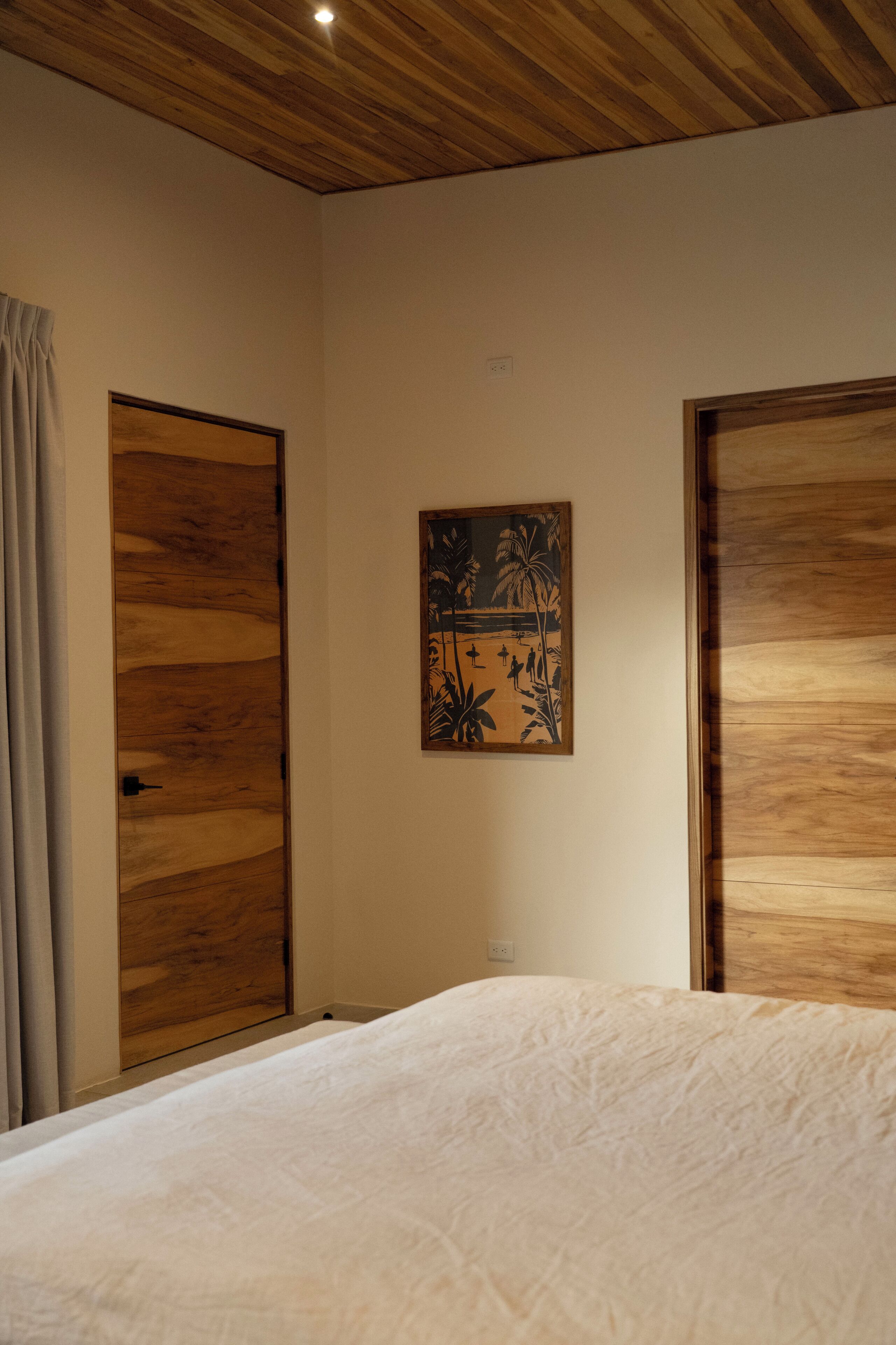 Un dormitorio con puertas de madera, una cama con colcha blanca y una obra de arte en la pared que representa una escena tropical.