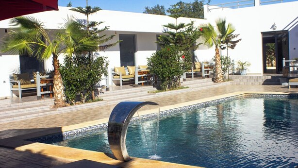A swimming pool with a waterfall feature, surrounded by palm trees and lounge chairs. - Dar naima  (Essaouira)