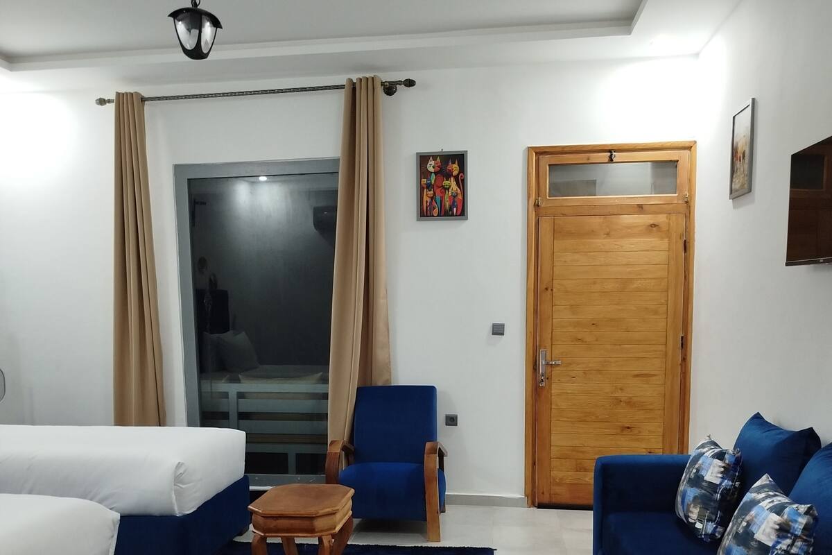 A modern hotel room with a large bed, a blue sofa, a blue armchair, a wooden coffee table, and a wooden door.