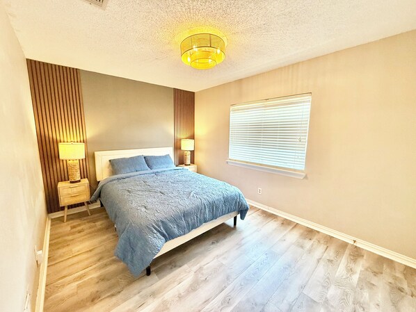 A bedroom with a bed, two bedside tables with lamps, a window with blinds, and a textured ceiling light.