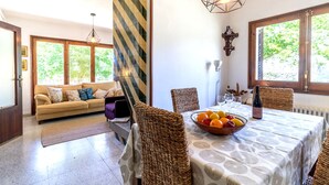 A dining area with a table set for six, a bowl of fruit, and a bottle of wine. There is a sofa with cushions in the background and a window with a view of greenery.