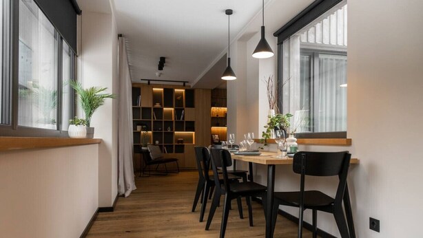 Un espace de salle à manger moderne avec une table et des chaises en bois, des luminaires suspendus et une vue sur la ville grâce à de grandes fenêtres.