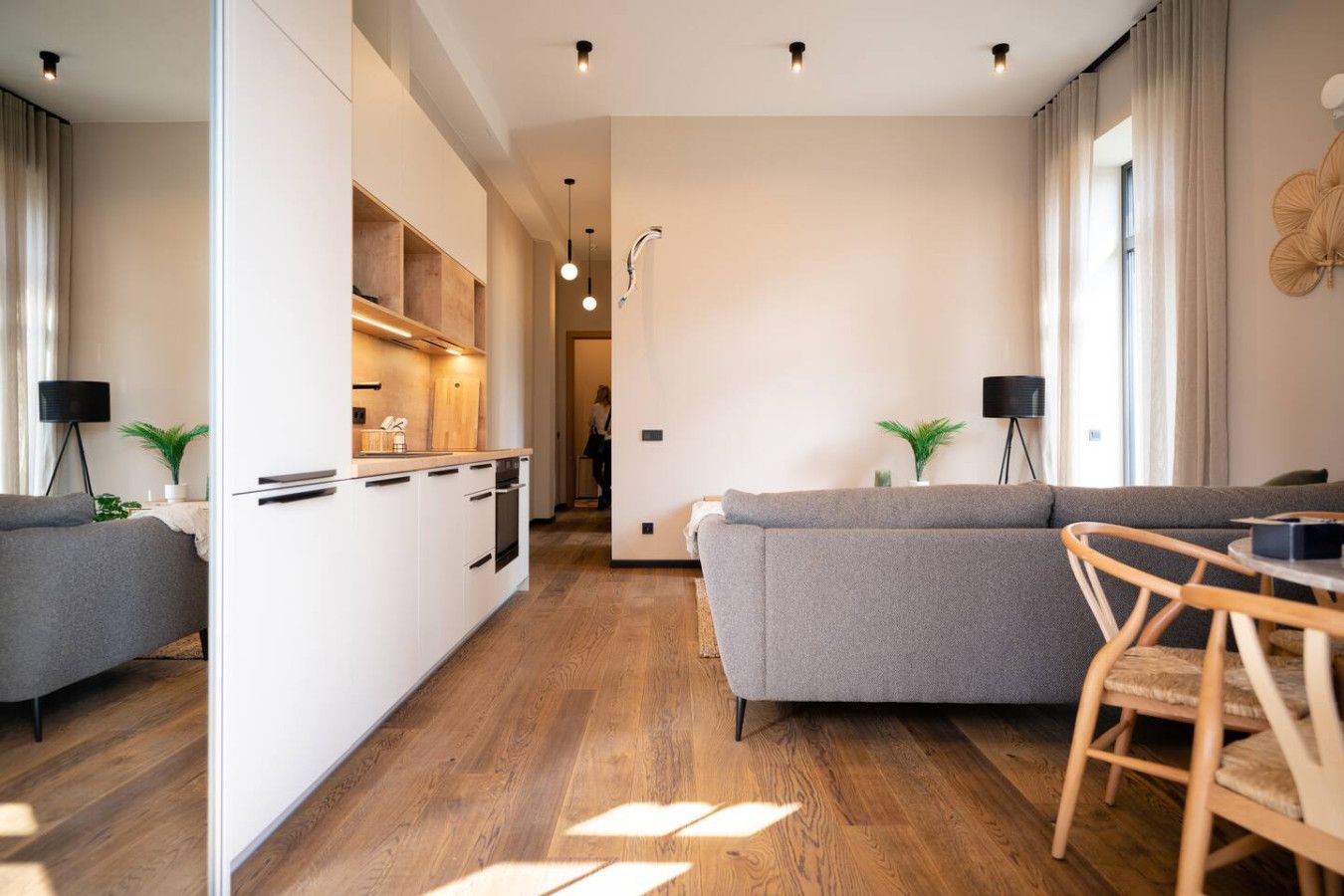 Una sala moderna con un sofá gris, una mesa de comedor de madera y una cocina con armarios blancos y horno empotrado.