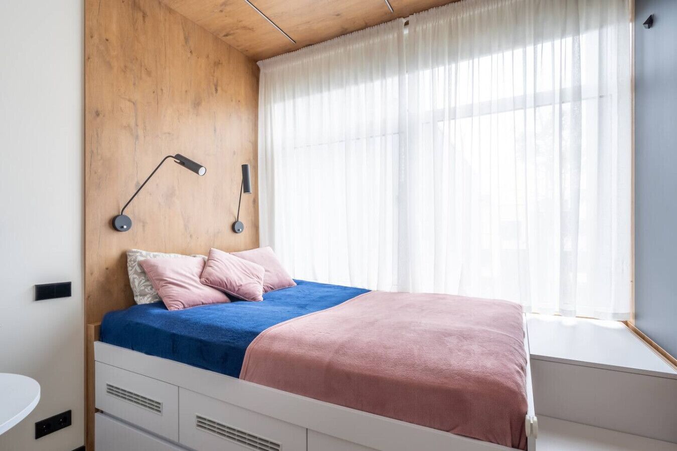 Une chambre à coucher avec une tête de lit en bois, un lit avec une literie bleue et rose, une table de chevet et une fenêtre ornée de rideaux légers.