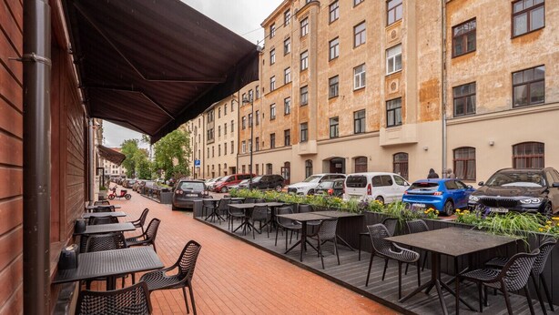 En udendørs café med borde og stole, et overdækket område og parkerede biler langs en gade med bygninger i baggrunden.