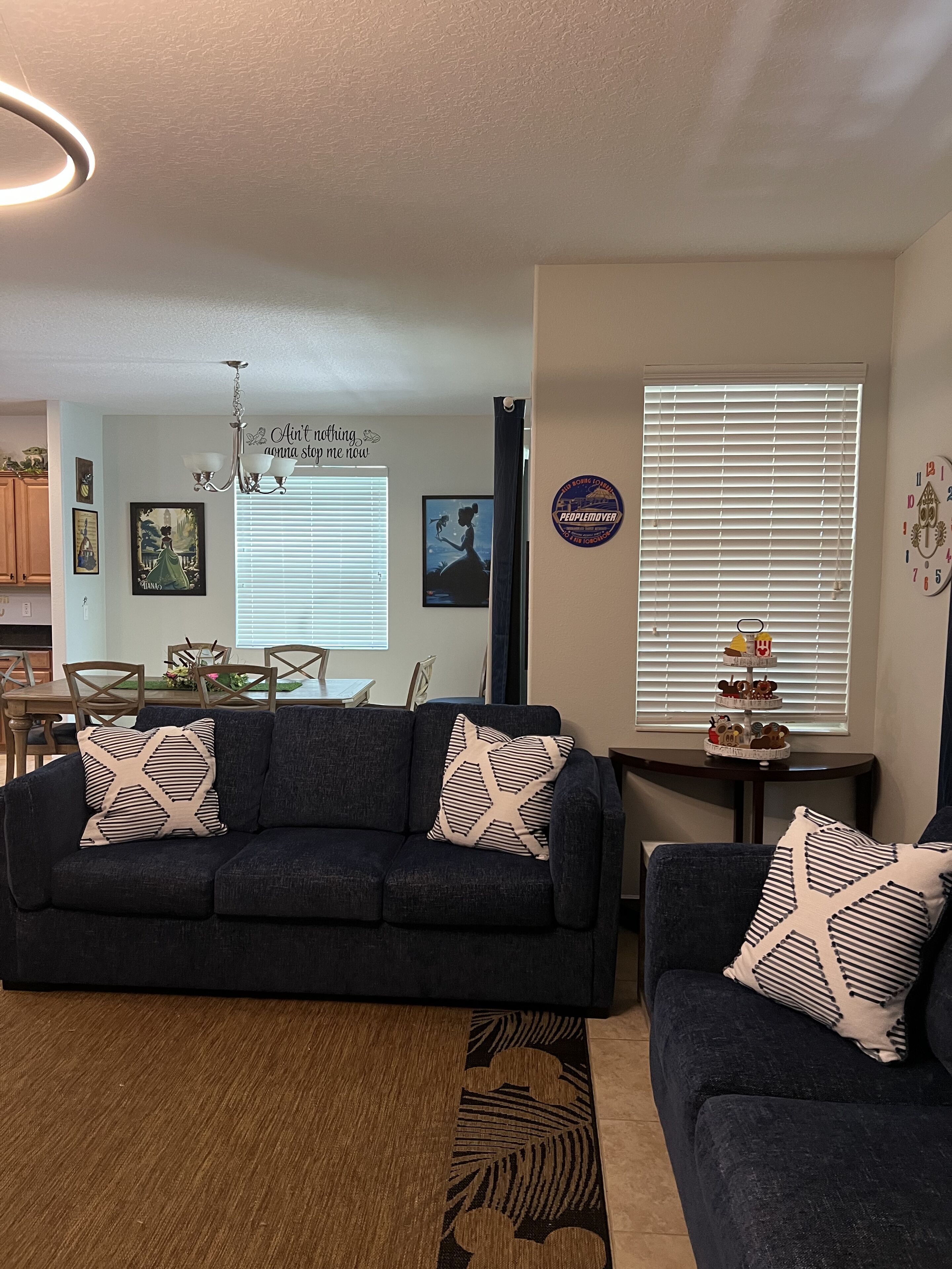 Ein Wohnzimmer mit einem dunkelblauen Sofa, einem kleineren Sofa und einem Couchtisch. Auf dem größeren Sofa liegen zwei Zierkissen. Eine Pendelleuchte hängt von der Decke, und eine Wanduhr ist sichtbar.