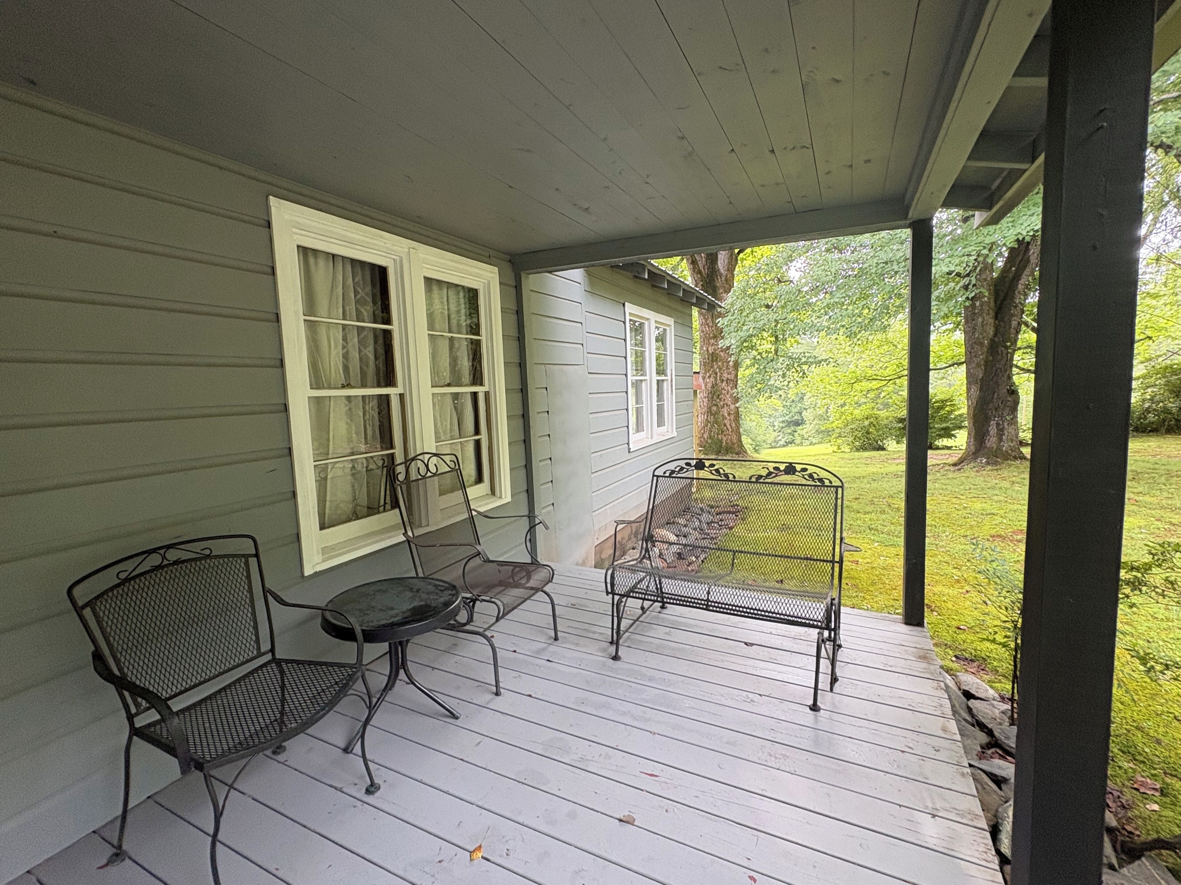 A covered porch with two metal chairs and a small table, a white door, and a view of a green landscape.
