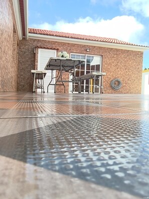 A covered outdoor area with a metal table and chairs, a stone wall, and a building with a tiled roof in the background. - Herdade das Hortênsias (São Jorge)