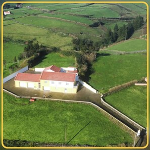 A large, single-story building with a red roof, surrounded by green fields and a stone wall. - Herdade das Hortênsias (São Jorge)