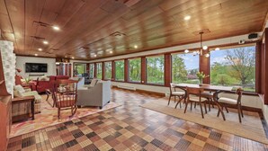 A spacious living room with a large TV, comfortable seating, a dining area, and large windows offering a view of the outdoors. - Friendly Hills | Passive Home • EV Charger (Pittsfield)