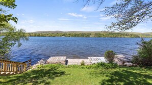 A lakeside deck with a wooden railing, a stone platform, and a view of a calm lake surrounded by greenery and hills. - Stockbridge Landings | 3 Mins To Tanglewood (Stockbridge)