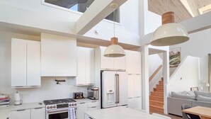 A modern kitchen with white cabinetry, a built-in oven, a stove, a refrigerator, and a large island. There is a staircase leading to a lofted area with a view of the outdoors.
