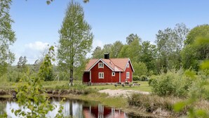 A red wooden house with a sloped roof, situated by a calm body of water, surrounded by lush greenery and trees. - Vacation home Bygget by Interhome (Simlångsdalen)