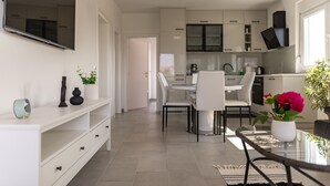 A modern kitchen with white cabinets, a dining area with a glass table, and a TV mounted on the wall.