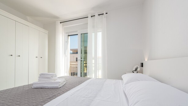 A modern bedroom with a large bed, white curtains, and a view of a building through a glass door.