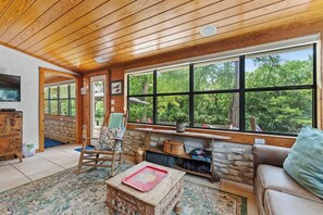 A cozy cabin interior with a stone fireplace, wooden ceiling, and large windows overlooking a forest.