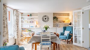 A dining area with a table set for four, surrounded by chairs, a clock on the wall, and shelves with various items.