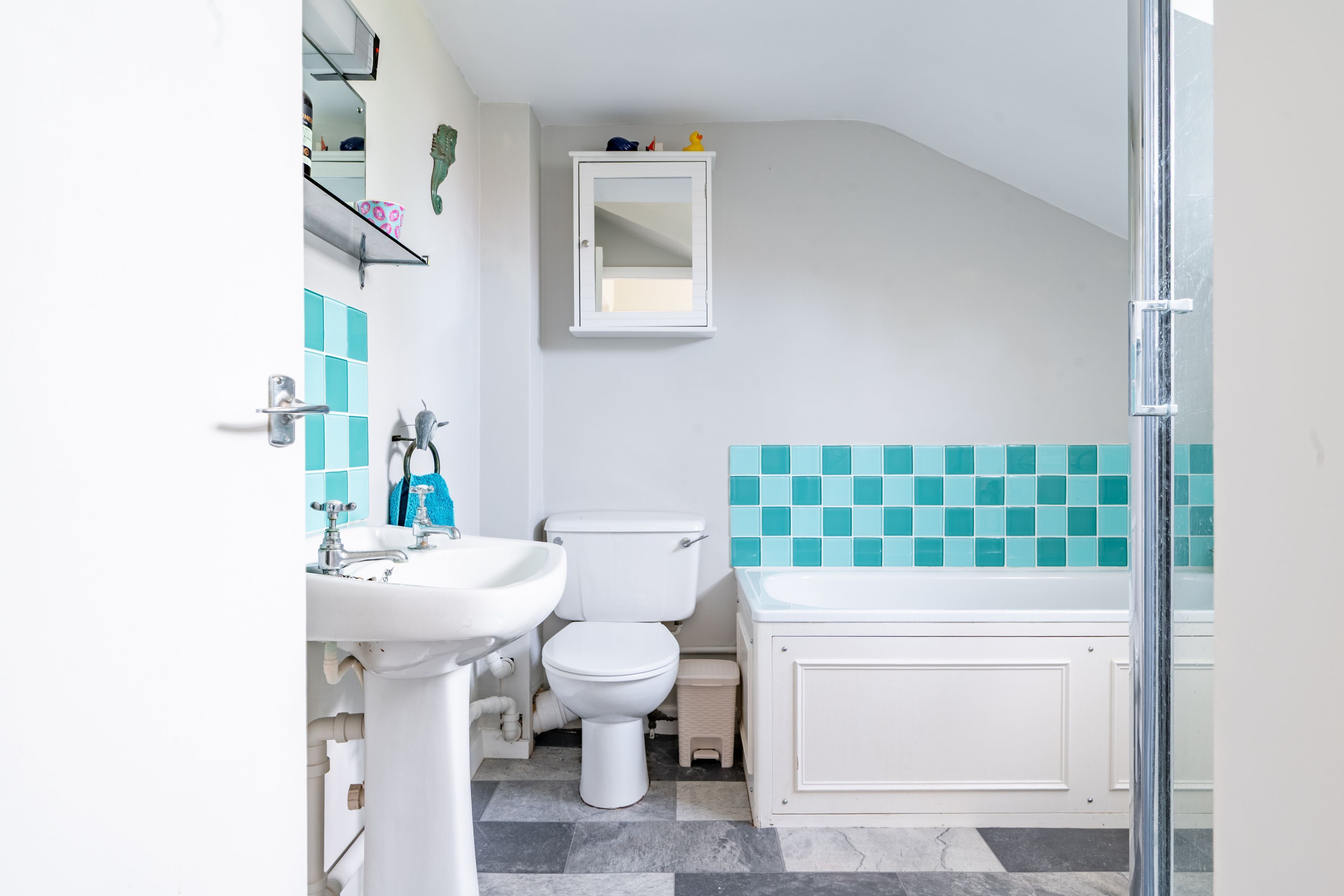Ein Badezimmer mit weißem Waschbecken, WC und Badewanne. Über dem WC befindet sich ein Spiegel und eine Ablage mit Toilettenartikeln. Der Boden ist mit einem gemusterten Fliesenmuster versehen.