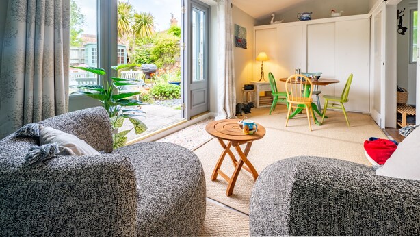 A cozy living room with a large sliding glass door leading to a balcony, a small round table with a lamp, and colorful chairs.