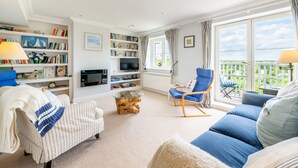 A living room with a sofa, armchair, and a television. There are bookshelves and a window with curtains.