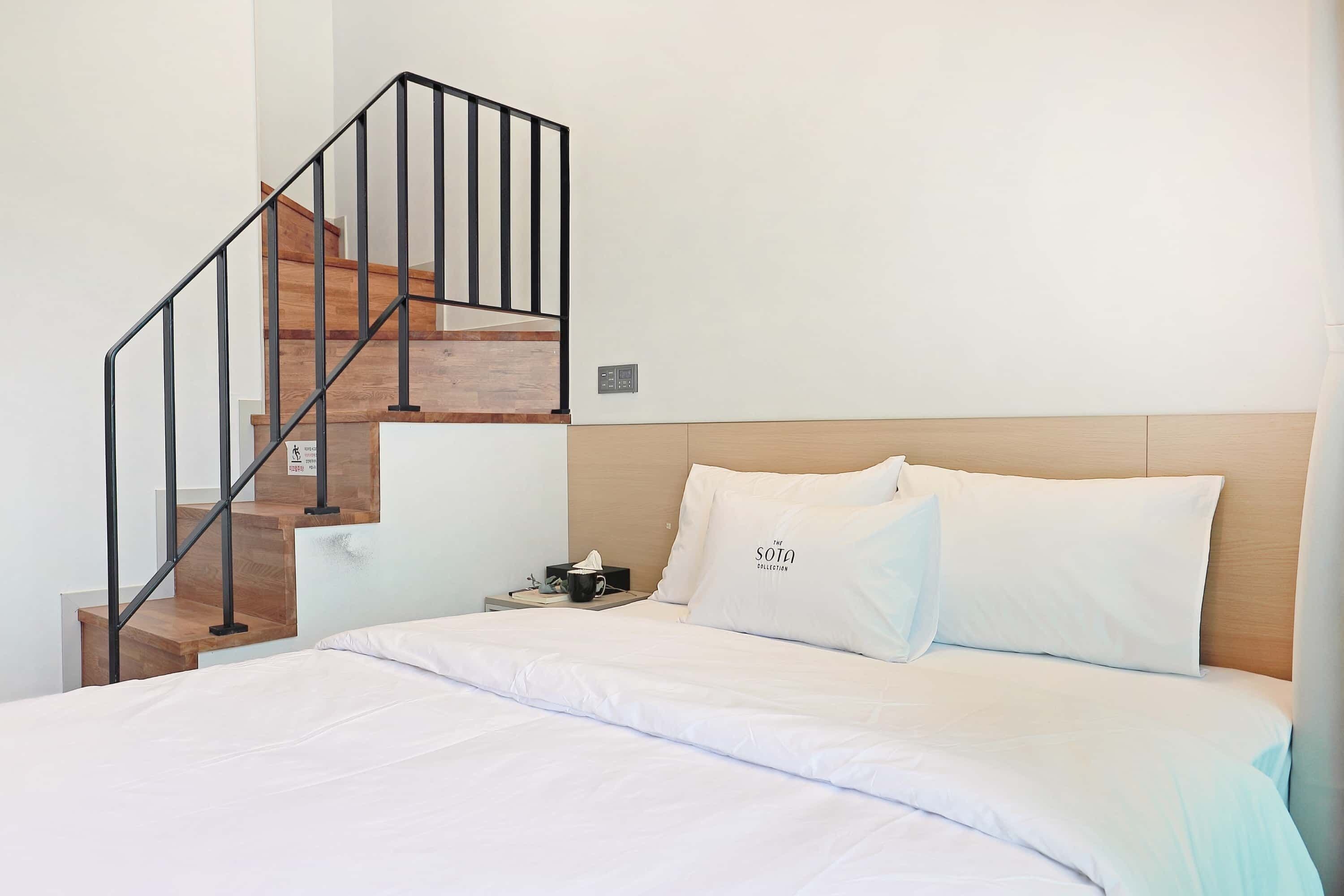A bedroom with a wooden staircase, a bed with white bedding, and a nightstand with a tissue box and a phone.