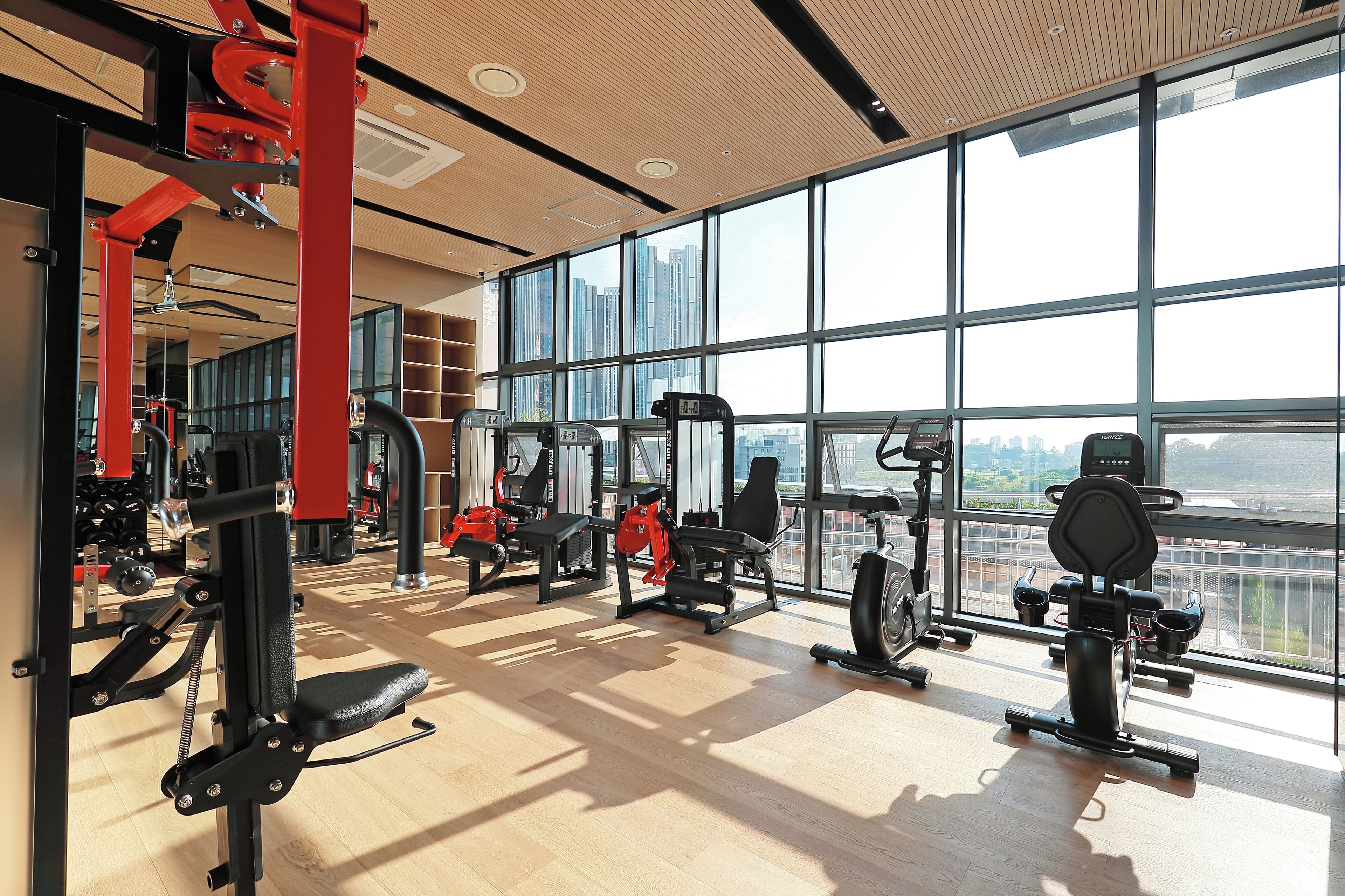 Une salle de sport spacieuse, dotée de grandes fenêtres, de divers appareils d’exercice et d’un plancher en bois.