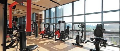 Une salle de sport spacieuse, dotée de grandes fenêtres, de divers appareils d’exercice et d’un plancher en bois.