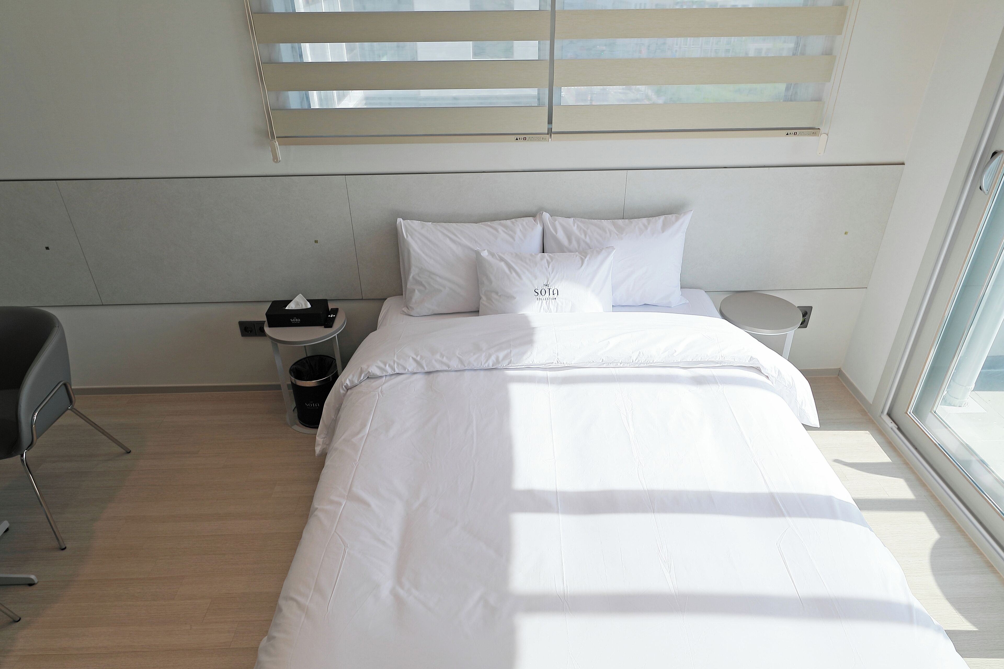 A neatly made bed with white linens, a bedside table with a lamp, and a window with blinds.
